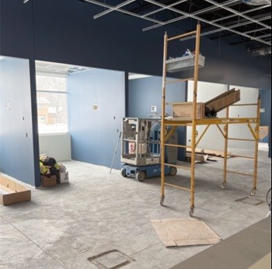 Construction area of a new building with small openings shows blue walls and open windows with blue paint and a yellow painting dolly in the middle on new concrete floors that are dusty