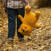 small child in blue jeans and plaid shirt holding teddy bear upside down on leaf covered path