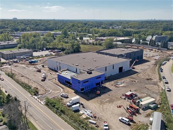 aerial image of white and gray building under construction in a dirt area with beams in front for small entry way with green trees and red brick building on the horizon 
