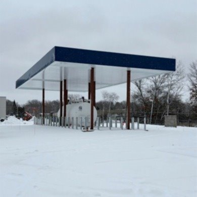 white fuel tank covered by a structure that has a blue flat roof with white on the interior with copper/brown beans supporting with snow on the ground