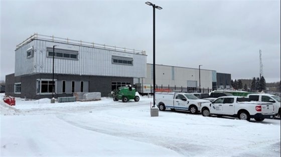 Gray and white building under construction in the winter with snow in parking area and several vehicles and white trucks in the lot