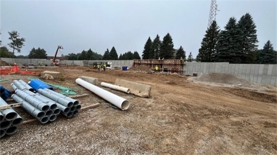 large dirt area with gray concrete wall on outside of the area surrounded by green trees with beams and barrels inside the enclosed area
