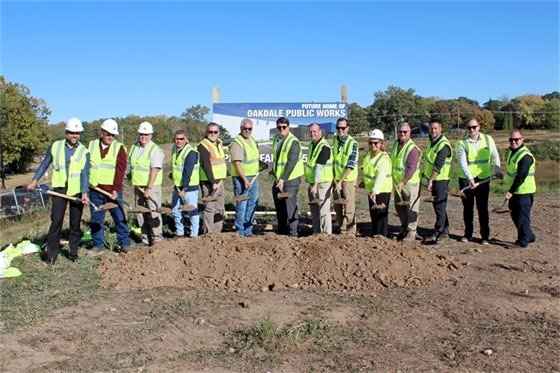 Group of 14 people in yellow construction vests holding shovels with dirt stand behind mound of dirt in front of large sign that reads "FUTURE HOME OF OAKDALE PUBLIC WORKS"