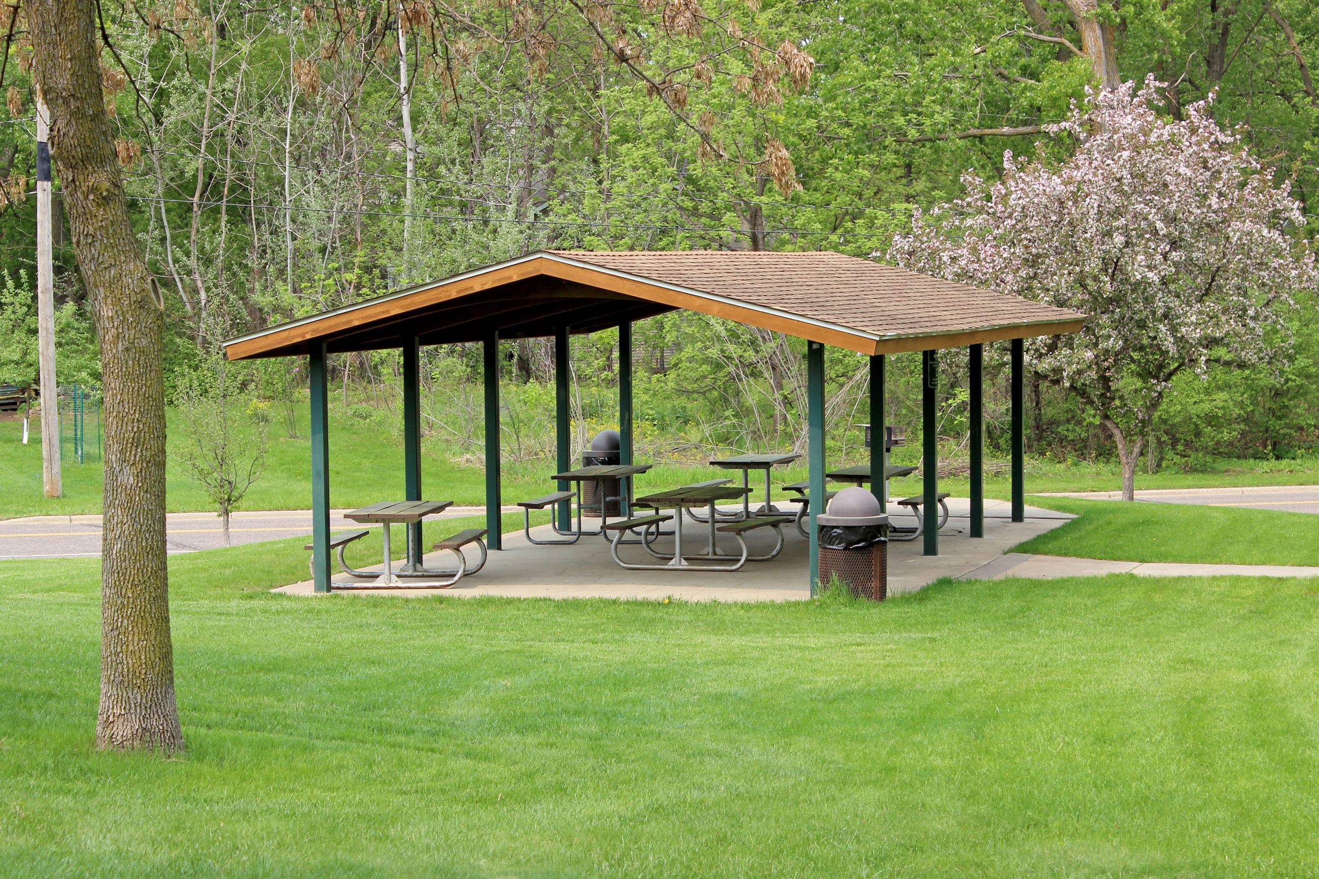 Picnic shelter with brown roof and picnic tables with green grass and pink tree