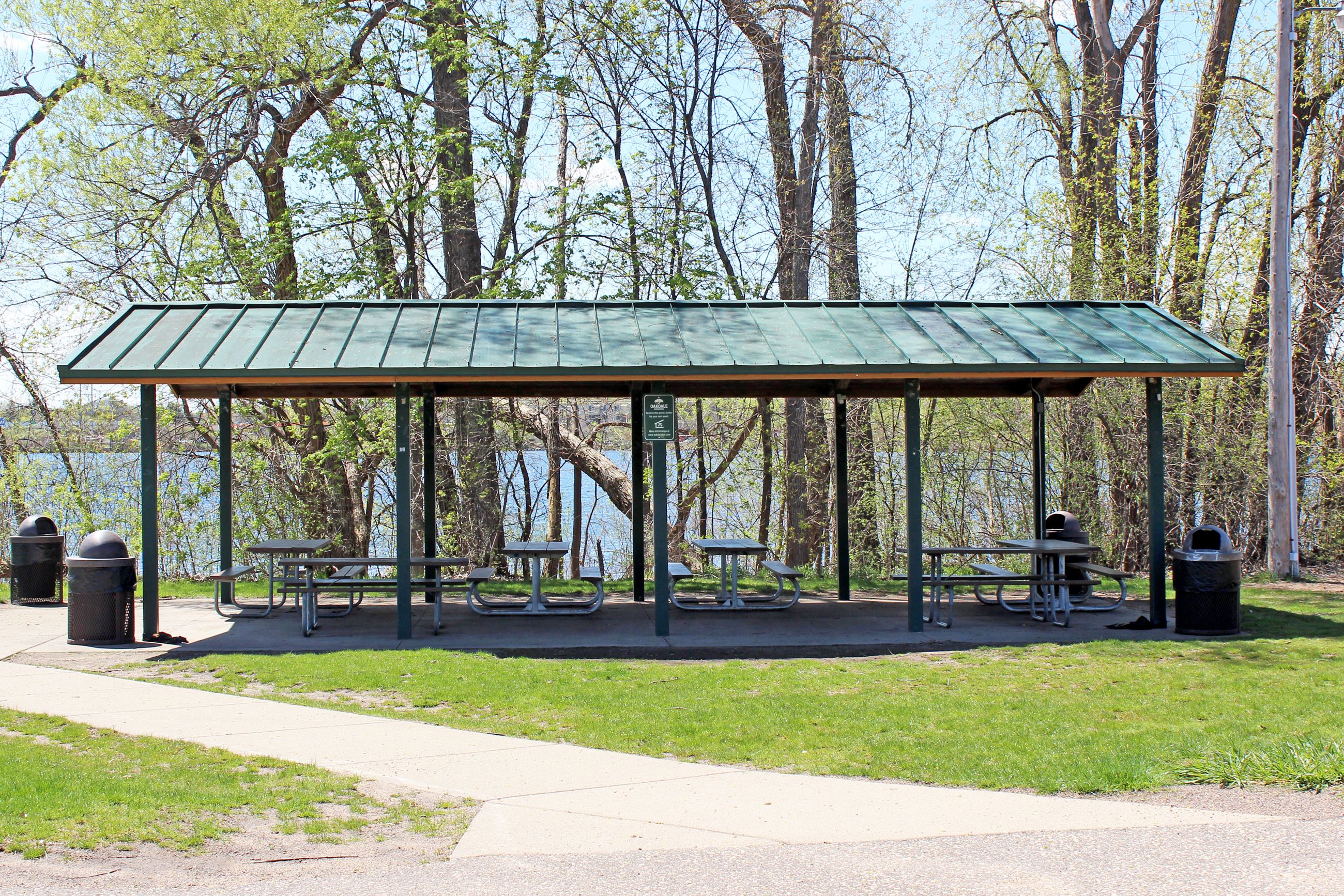 Green picnic shelter with tables and garbage cans in shadows of trees with lake