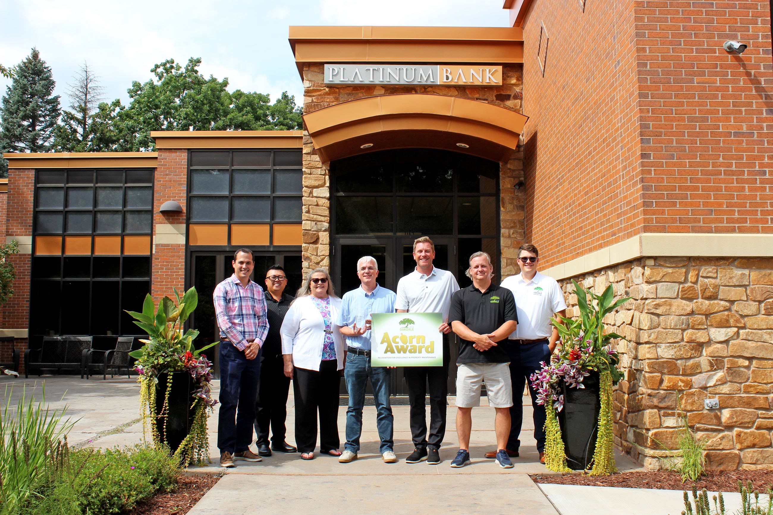 group of seven people pose holding a sign that reads "Acorn Award" in front of orange brick bu