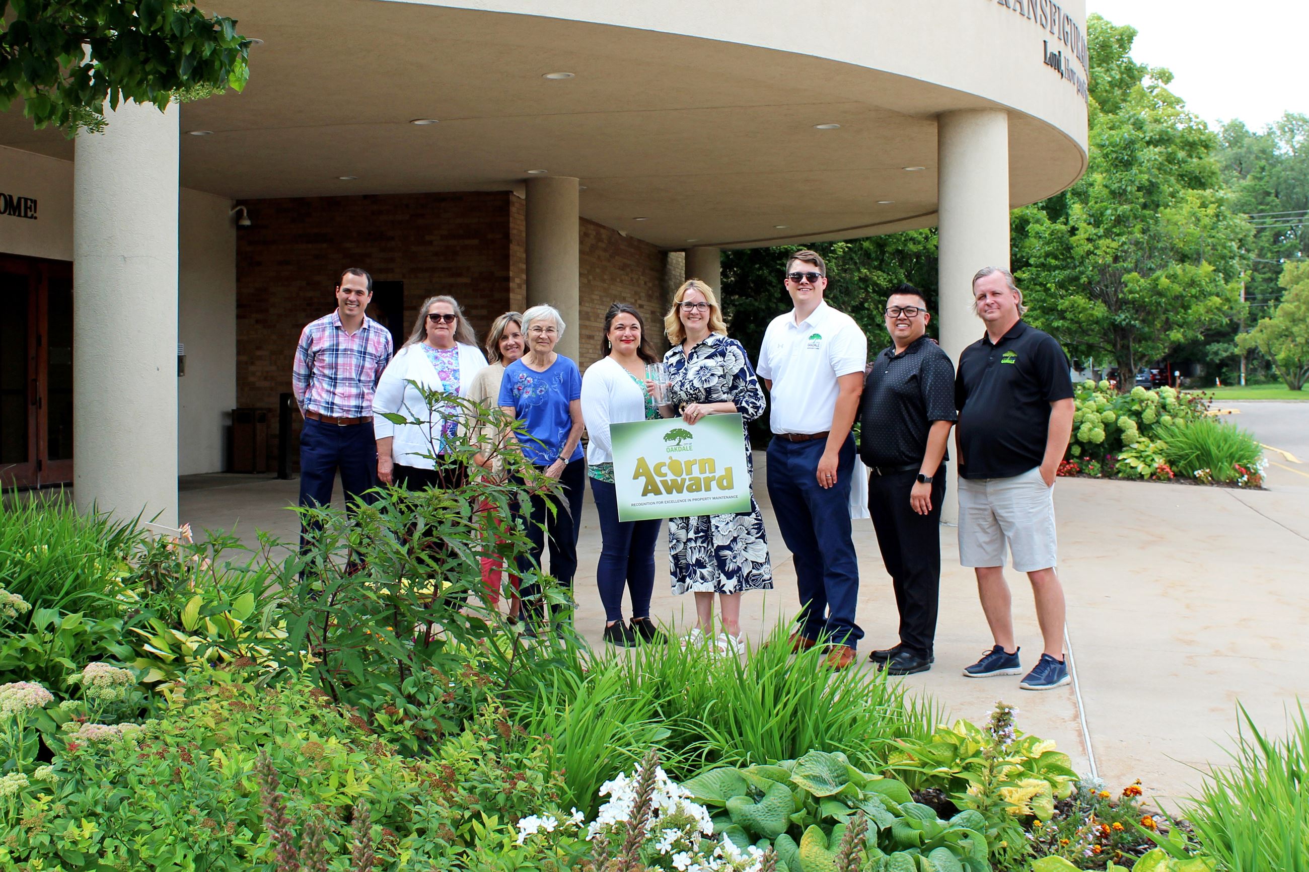 Group of nine people holding a sign that reads "Acorn Award" pose for photo in front of tan bu
